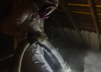 A worker in protective gear sprays insulation material in an attic under a wooden roof.