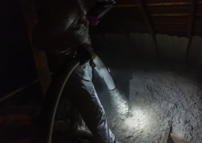 Person in protective suit spraying insulation material in a dimly lit attic with a hose.