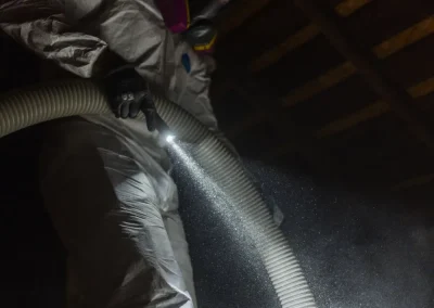 Person in protective suit spraying insulation or dust in a dark attic, with light highlighting airborne particles.