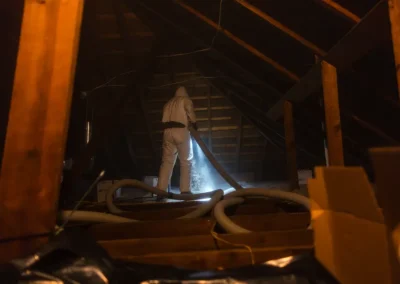 Person in protective suit spraying insulation in a dimly lit attic with boxes and hoses visible.