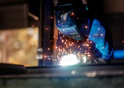 A person wearing protective gear welds metal, with bright sparks flying in a dark workshop.