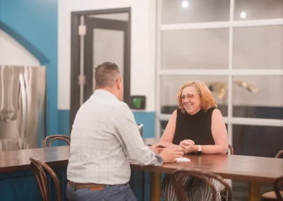 Two people sit at a table in a modern office, smiling and having a friendly conversation.