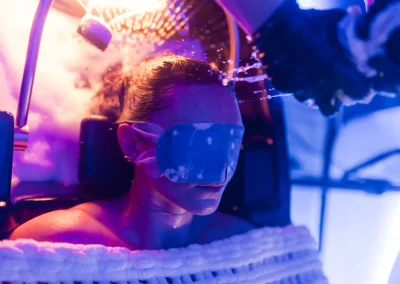 Person wearing eye mask receives cold therapy with mist under purple and blue light in a spa setting.