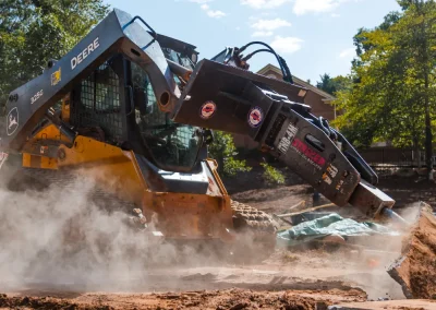 A John Deere skid steer uses a hydraulic breaker attachment to break concrete, creating dust.