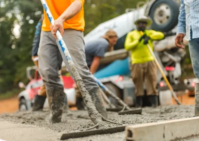 Workers spread wet concrete on a construction site, with a cement mixer truck in the background.