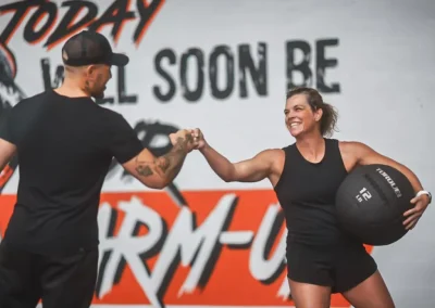 Two people in workout gear fist bump; one holds a medicine ball and smiles. Gym mural is in the background.