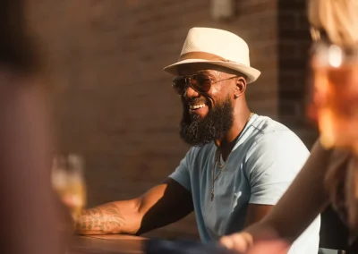 Man in a hat and sunglasses smiles while sitting outdoors at a table with others in sunlight.