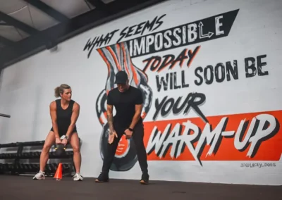 A woman lifts a kettlebell while a trainer watches in a gym with a motivational wall mural.