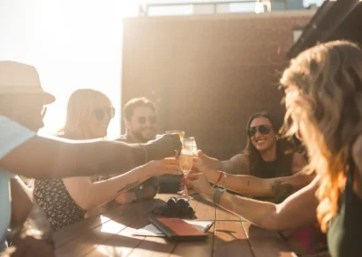 Five friends clink glasses at an outdoor table, smiling in the sunlight.