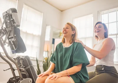 Two women smile during a seated massage session in a bright room with large windows.