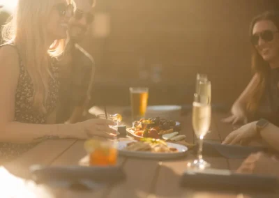 Four people sit at a sunlit outdoor table with drinks and food, smiling and talking together.