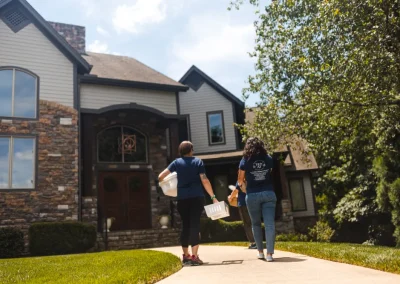 Two people carrying boxes walk toward a large stone house on a sunny day with green trees and grass.