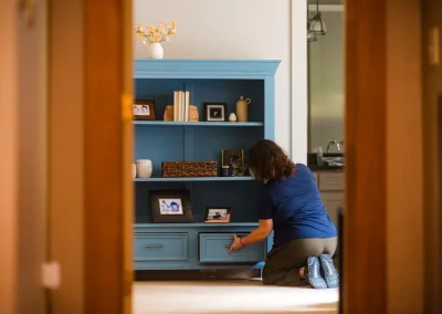 Person kneeling and organizing a blue cabinet with photos and decor, seen through a doorway.