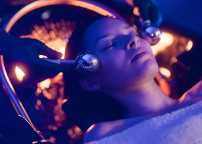 Person receiving a facial massage with metal rollers in a dim, blue-lit spa setting.