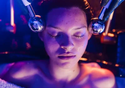 Woman with closed eyes receives a facial treatment using metal rollers under blue and purple lighting.