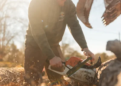 Man in a cap cuts a log with a chainsaw outdoors on a sunny day, wood chips flying.