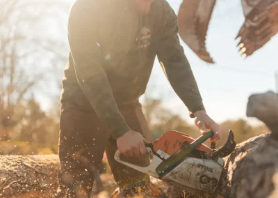 Man wearing cap cuts a log with a chainsaw outdoors on a sunny day, wood chips flying.