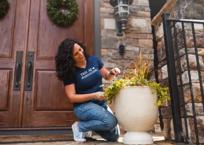 Woman kneeling and tending to plants in a large white pot outside a stone house with double wooden doors.