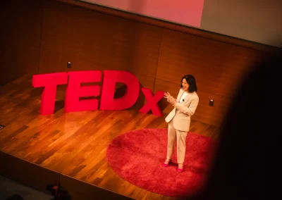 A woman in a white suit speaks on stage beside a large red TEDx sign and a round red carpet.