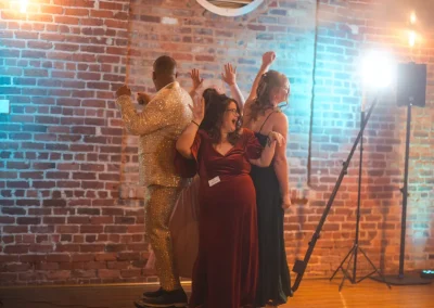 Four people in formal attire dance enthusiastically together in front of a brick wall and bright spotlight.