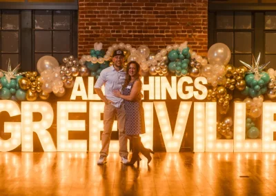 A couple poses in front of large lit “GREENVILLE” letters with balloon decorations and brick walls.