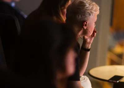 People sit in a dimly lit room, focusing on something out of view, with a phone on a small table nearby.
