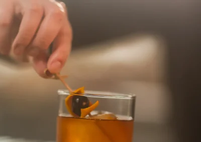 Hand placing a cocktail pick with an orange twist and cherry into a glass of amber-colored drink on a table.