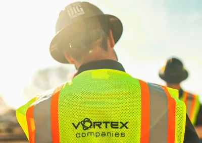 Worker in neon safety vest and hard hat stands outdoors, sunlight shining, “Vortex Companies” logo visible.