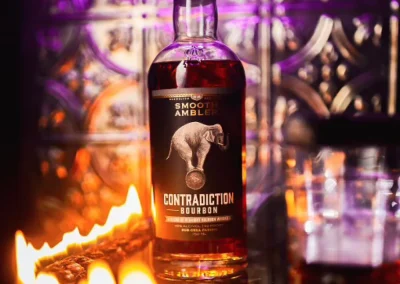 A bottle of Contradiction Bourbon stands on a reflective surface with flames and purple lighting in the background.