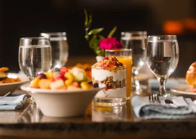 A breakfast table with fruit salad, yogurt parfait, water glasses, and a flower vase in the center.