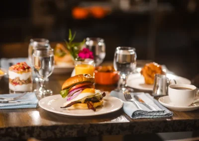 Burger with cheese, bacon, and veggies on a plate with drinks, coffee, and desserts on a restaurant table.