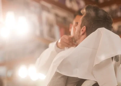 A man sits in a barber chair, covered with a cape, while a barber trims his beard in a shop.