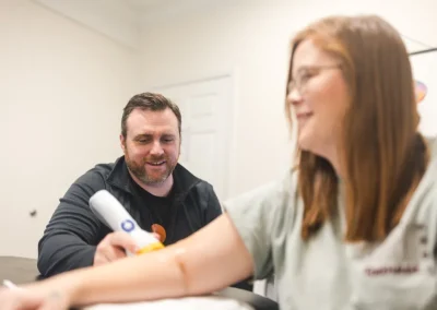 A man holds a medical device to a woman’s arm while she smiles in a bright room.