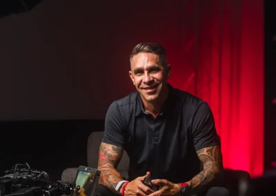 Man with tattoos sits on armchair, smiling at camera, with photography equipment in foreground, red lighting behind.