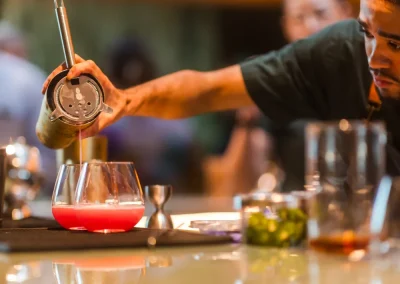 Bartender pouring a red cocktail into a glass at a bar, bottles blurred in the background.