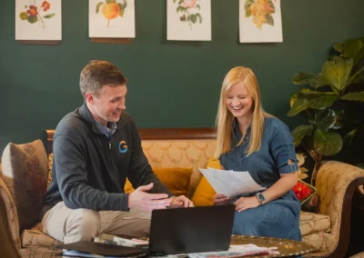 Two people sit on a couch, smiling and discussing papers, with a laptop on the table in front.