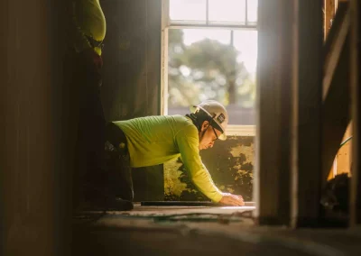 Two construction workers in hard hats work near a window, one kneeling and inspecting the floor.