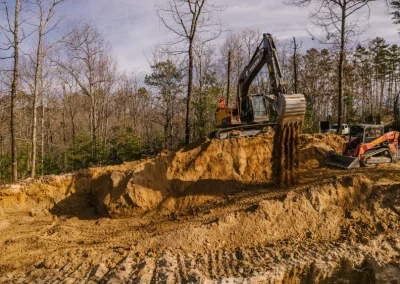Excavator pours dirt into a pit beside a small loader in a forested, cleared construction site.