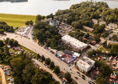 Aerial view of a crowded outdoor event with tents, parked cars, and nearby lake surrounded by trees.
