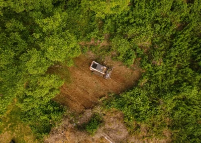 A bulldozer clears land in a forested area, seen from above, surrounded by dense green trees.