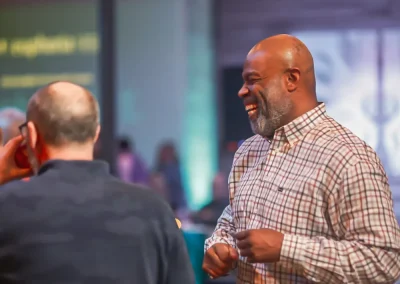 Two men talking and smiling at an indoor event with colorful lights in the background.