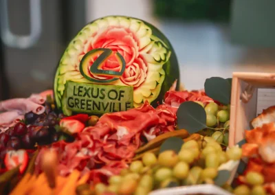 Carved watermelon with “Lexus of Greenville” among meats, grapes, and snacks on a platter.