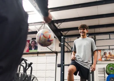 Teen boy in a gym smiles and lifts his leg as a soccer ball is tossed toward him by another person.
