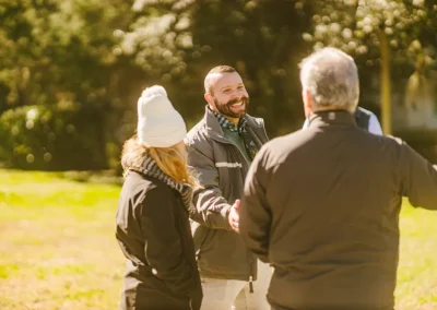 Smiling man in jacket shakes hands with another person outdoors, with trees and sunlight in the background.