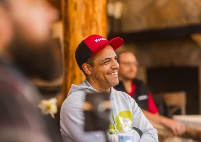 Man in a red cap smiling, sitting at a table indoors with others in a cozy, rustic setting.