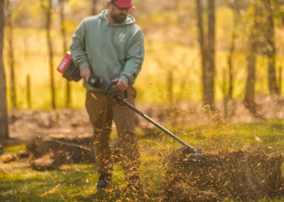 Person using a string trimmer to cut dry grass, with debris flying up, in a sunlit outdoor area.