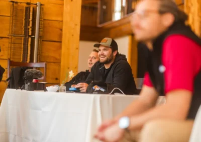 Three people sit at a table indoors, one smiling, with wooden walls and equipment in the background.