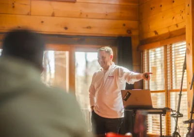 Smiling man in white shirt stands by a laptop, pointing while giving a presentation in a wooden room.