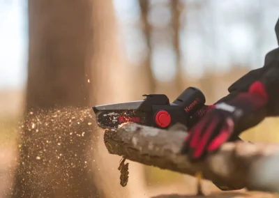 Close-up of gloved hands using an electric chainsaw to cut a tree branch, wood chips flying.