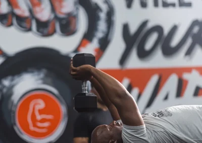 Man lying on a bench, lifting a dumbbell overhead in a gym with bold mural on the wall behind him.